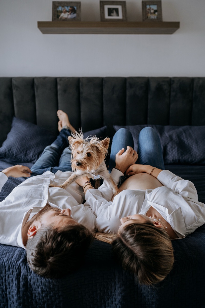 Pareja recostada en cama con su perro, sesión de embarazo lifestyle