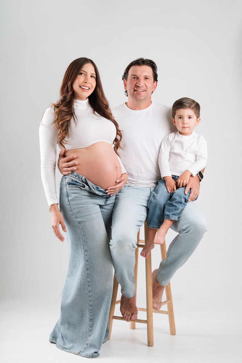 Familia en sesión de embarazo: mamá, papá y su hijo pequeño sentados en un banco, fondo blanco